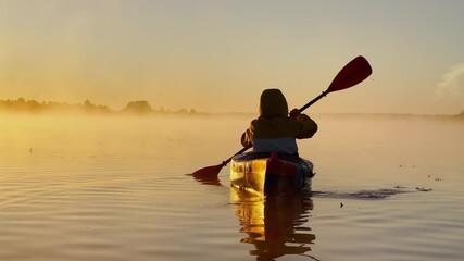 Wall Mural - Kayaker floats on calm water through the morning fog above the water at sunrise, the silhouette of a man with a paddle on a kayak, the golden color of the water, warm water and cold air, magic light