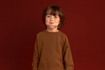 Funny picture of handsome boy with stylish haircut in sweater blowing cheeks, having fun, making faces, fooling around, looking at camera ready to laugh out loud. Happy carefree childhood