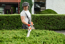 Handsome Caucasian Man In Uniform, Face Defender And Protective Gloves Cutting Overgrown Bushes On Backyard. Competent Gardener Using Petrol Hedge Trimmer For Work Outdoors.