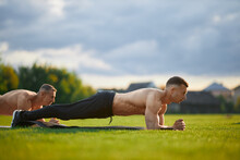 Athletic Shirtless Men Doing Plank Exercise On Yoga Mat On Outdoors Stadium. Two Caucasus Bodybuilders Having Intense Workout On Fresh Air. Strength And Endurance Concept.