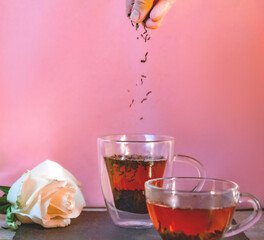 Two glass cups of tea on a plain pinc background. The rose lies on the table. Hand pouring tea. Front view