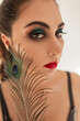 Creative portrait of a pretty young fashion vogue girl with bright makeup and a peacock feather looking at the camera. Beautiful female brown eyes