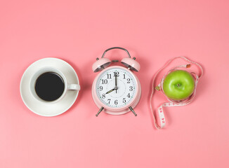 flat lay of pink vintage alarm clock, black coffee cup, green apple and measuring tape on pink background. Morning routine and weight loss concept.