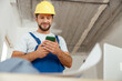 © Kostiantyn - Focused repairman in workwear and hardhat smiling while using smartphone, standing indoors during renovation work