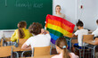 Female teacher holding rainbow-colored LGBT community flag and explaining it meaning for kids during lesson dedicated to family love.