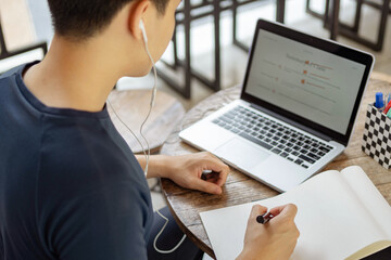 Online studying concept a black-haired student doing his homework by searching information on the internet and using this black pen to write the conclusion of his research on the paper