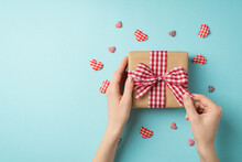 First Person Top View Photo Of St Valentine's Day Decor Hands Untying Checkered Ribbon Bow On Kraft Paper Giftbox Surrounded By Decorative Hearts On Isolated Pastel Blue Background With Empty Space