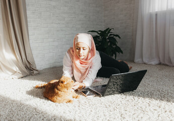 Wall Mural - young modern Muslim woman in hijab sits on the floor and works on a laptop at home, online training, remote work.