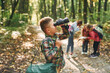 © standret - Boy with binoculars standing in front of his friends. Kids in green forest at summer daytime together