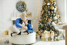 A Young Family - Mom And Son, Sitting In A Bright Christmas Decorated Room In Gold Tones And Tenderly Embracing, Holding A Present With A Live White Rabbit In Their Hands.