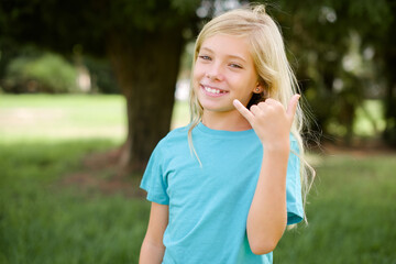 Wall Mural - Caucasian little kid girl wearing blue T-shirt standing outdoors smiling doing phone gesture with hand and fingers like talking on the telephone. Communicating concepts.