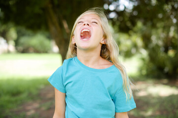 Caucasian little kid girl wearing blue T-shirt standing outdoors angry and mad screaming frustrated and furious, shouting with anger. Rage and aggressive concept.