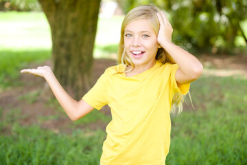 Shocked amazed surprised beautiful Caucasian little kid girl wearing yellow T-shirt standing outdoors hold hand offering proposition