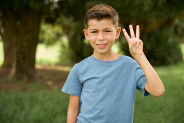 Wall Mural - beautiful Caucasian little kid boy wearing blue T-shirt standing outdoors showing and pointing up with fingers number three while smiling confident and happy.