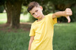 Caucasian little kid boy wearing yellow T-shirt standing outdoor  feeling angry, annoyed, disappointed or displeased, showing thumbs down with a serious look