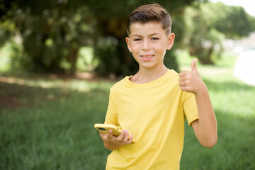 Wall Mural - Portrait of Caucasian little kid boy wearing yellow T-shirt standing outdoors using and texting with smartphone  happy with big smile doing ok sign, thumb up with fingers, excellent sign