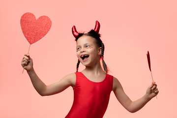 Wall Mural - Little happy girl with pigtails in costume of little devil holding two red hearts in hands against pink background