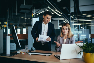 Wall Mural - Two employees in a modern office, an Asian man and a woman working at a table, colleagues discussing and consulting, thinking about a joint project