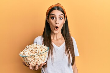 Wall Mural - Beautiful brunette young woman holding popcorn scared and amazed with open mouth for surprise, disbelief face