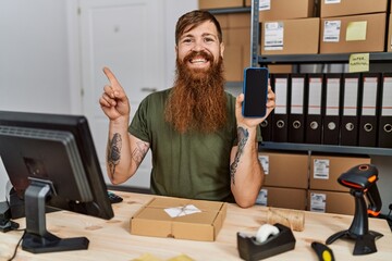 Wall Mural - Redhead man with long beard working at small business holding smartphone smiling happy pointing with hand and finger to the side