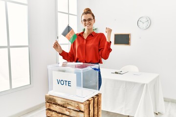 Poster - Young brunette woman by ballot box holding afghanistan flag screaming proud, celebrating victory and success very excited with raised arms