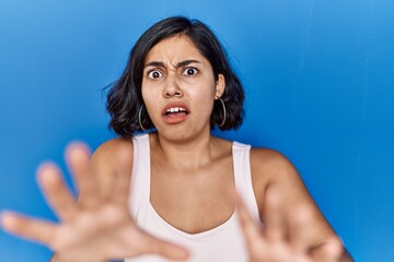 Canvas Print - Young hispanic woman standing over blue background afraid and terrified with fear expression stop gesture with hands, shouting in shock. panic concept.