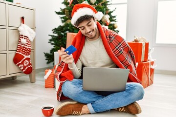 Canvas Print - Young hispanic man wearing christmas hat using laptop and credit card at home.