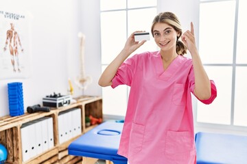 Poster - Young physiotherapist woman working at pain recovery clinic holding credit card surprised with an idea or question pointing finger with happy face, number one