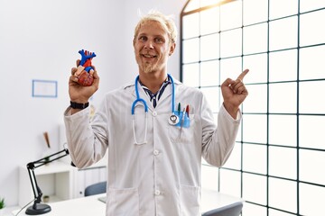 Canvas Print - Young blond man wearing doctor uniform holding heart at clinic smiling happy pointing with hand and finger to the side