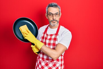 Poster - Handsome middle age man with grey hair wearing apron holding scourer washing pan relaxed with serious expression on face. simple and natural looking at the camera.