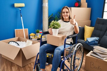 Poster - Young woman sitting on wheelchair moving to a new home approving doing positive gesture with hand, thumbs up smiling and happy for success. winner gesture.