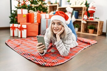 Poster - Young caucasian girl wearing christmas hat using smartphone lying on the floor at home.