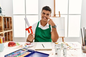 Poster - Young hispanic man at art studio holding degree pointing with hand finger to face and nose, smiling cheerful. beauty concept