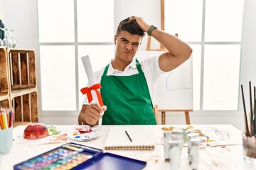 Sticker - Young hispanic man at art studio holding degree confuse and wondering about question. uncertain with doubt, thinking with hand on head. pensive concept.