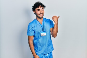 Wall Mural - Young hispanic man wearing blue male nurse uniform smiling with happy face looking and pointing to the side with thumb up.