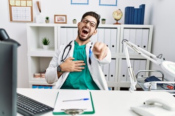 Poster - Young man with beard wearing doctor uniform and stethoscope at the clinic laughing at you, pointing finger to the camera with hand over body, shame expression