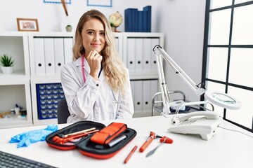 Poster - Young beautiful doctor woman with reflex hammer and medical instruments looking confident at the camera with smile with crossed arms and hand raised on chin. thinking positive.