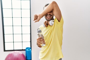 Sticker - Young hispanic man wearing sportswear and drinking water at the gym smiling cheerful playing peek a boo with hands showing face. surprised and exited