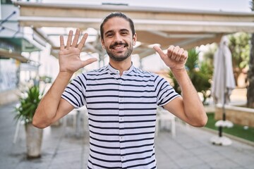 Wall Mural - Young hispanic man with beard outdoors at the city showing and pointing up with fingers number six while smiling confident and happy.