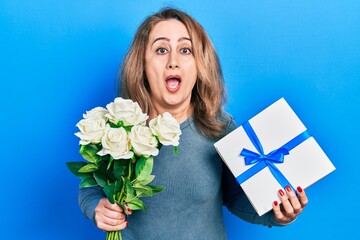 Poster - Middle age caucasian woman holding anniversary present and bouquet of flowers afraid and shocked with surprise and amazed expression, fear and excited face.