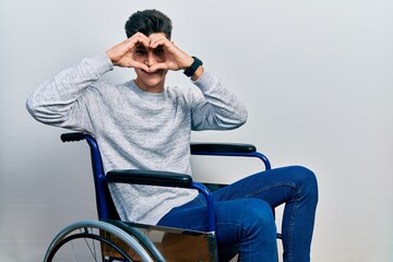 Wall Mural - Young hispanic man sitting on wheelchair doing heart shape with hand and fingers smiling looking through sign