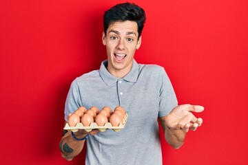 Young hispanic man showing fresh white eggs celebrating achievement with happy smile and winner expression with raised hand