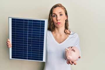 Poster - Young blonde woman holding photovoltaic solar panel and piggy bank depressed and worry for distress, crying angry and afraid. sad expression.