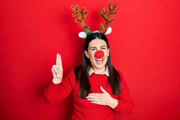 Wall Mural - Young hispanic woman wearing deer christmas hat and red nose smiling swearing with hand on chest and fingers up, making a loyalty promise oath