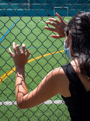 Young Brunette Woman Using a Blue Mask is Looking Through the Mesh of the Soccer Field