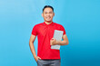 Portrait of smiling Asian handsome young man in red shirt holding laptop and looking at camera isolated on blue background