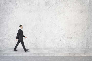 Poster - Side view of young businessman walking in concrete interior with mock up place on wall. Success concept.