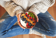 Healthy breakfast at home. Woman wearing jeans and sweater holding bowl with yogurt and corn flakes topped of  blueberry, raspberry, sliced kiwi and strawberry. Vegetarian food