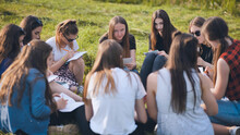 A Group Of Female Students Are Sitting In A Circle On A Meadow For Collective Work With Notebooks.