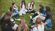 © Довидович Михаил - A group of female students are sitting in a circle on a meadow for collective work with notebooks.